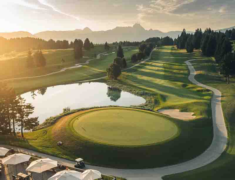 Luftaufnahme einer luxuriösen Golfanlage in Oberbayern mit einer Clubhaus-Terrasse im Vordergrund, einem kreisrunden Green am See und Blick auf die Chiemgauer Alpen im goldenen Abendlicht.