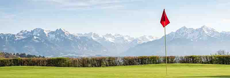 Gepflegtes grünes Golf-Green mit einer roten Fahne im Vordergrund und einem beeindruckenden Panorama der oberbayerischen Alpen unter blauem Himmel, ideal für einen Golfurlaub in Oberbayern.